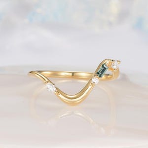 Peut inclure: Une bague dorée au design incurvé, ornée d'une pierre rectangulaire de couleur teal et de plusieurs petites pierres rondes et claires. La bague a une apparence délicate et élégante, adaptée à une occasion spéciale.
