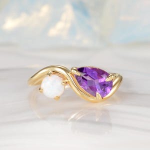 May include: A gold ring featuring a pear-shaped purple amethyst and a round white opal. The gemstones are set in a swirling gold band, creating a unique and elegant design. The ring is displayed on a reflective surface.