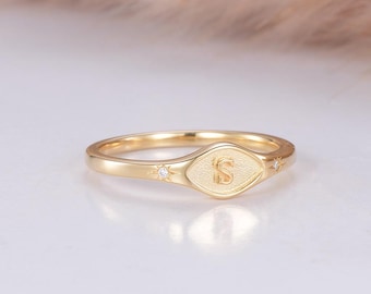 May include: A gold ring with a diamond-shaped center featuring the letter "S". The ring has a delicate band and small accent diamonds on each side of the center design. The ring is set against a soft, neutral background.