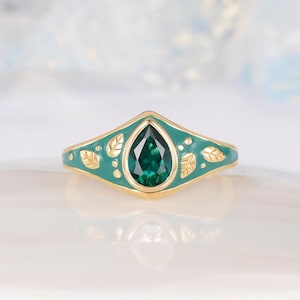 May include: A gold ring with a teardrop-shaped emerald green gemstone. The ring band is teal with gold leaf accents. The ring is a close-up shot, showcasing the intricate details of the jewelry.