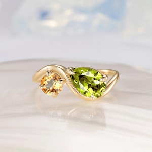 Peut inclure: Une bague en or ornée d'une pierre jaune taille ronde et d'une pierre verte taille poire. Les deux pierres sont serties dans une monture de style bypass qui s'enroule autour de chaque gemme, créant un design fluide et continu.
