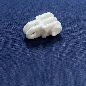 May include: A small, white 3D-printed object with a unique shape, possibly a component or decorative piece. The object has a textured surface and appears to be made of plastic. It sits on a blue fabric background.