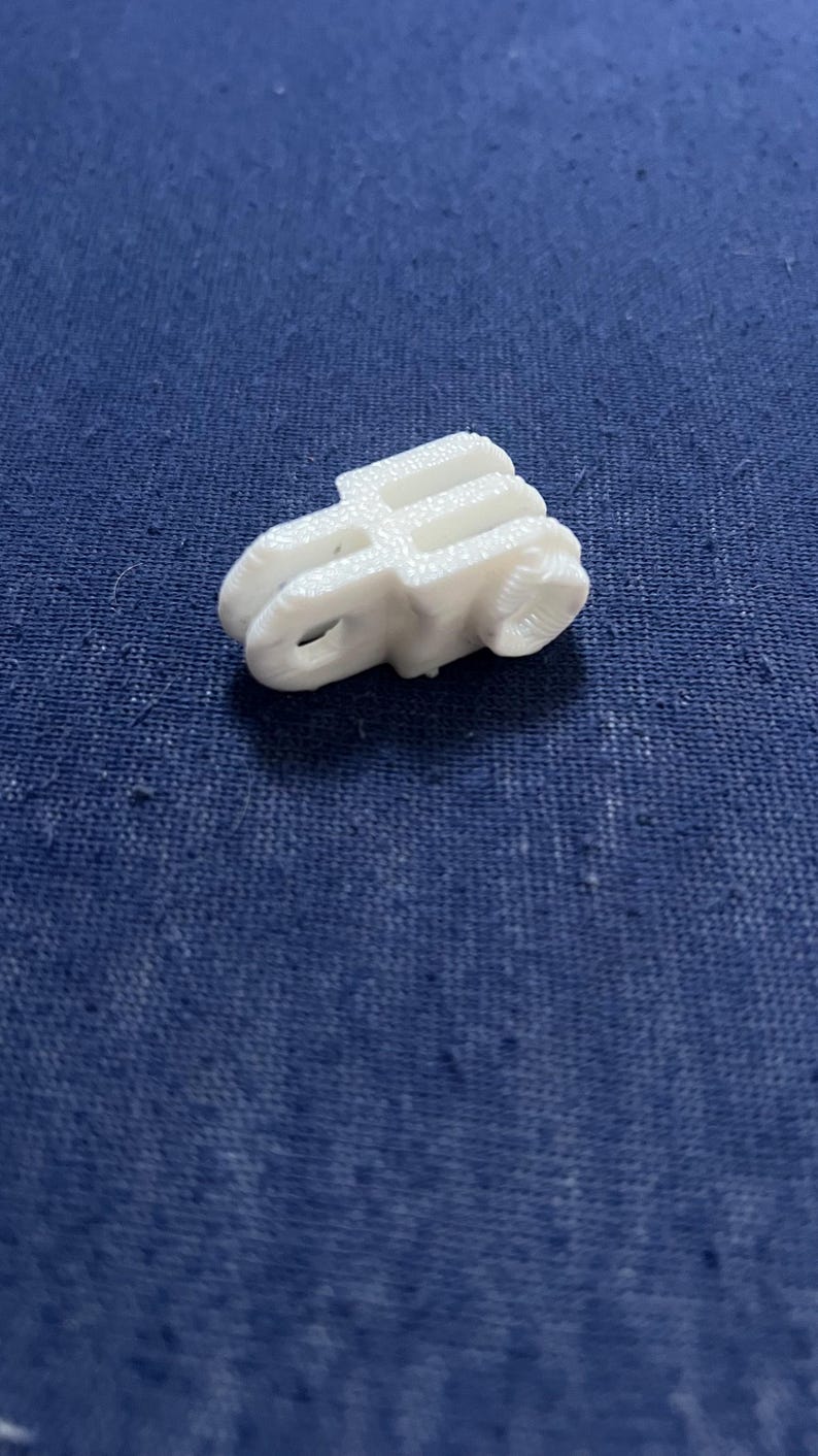 May include: A small, white 3D-printed object with a unique shape, possibly a component or decorative piece. The object has a textured surface and appears to be made of plastic. It sits on a blue fabric background.