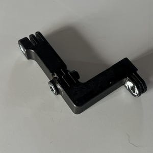 May include: A black, L-shaped metal bracket with two mounting holes and a slotted end. The bracket has a screw in the center and is designed for mounting or securing equipment. The object is on a white surface.