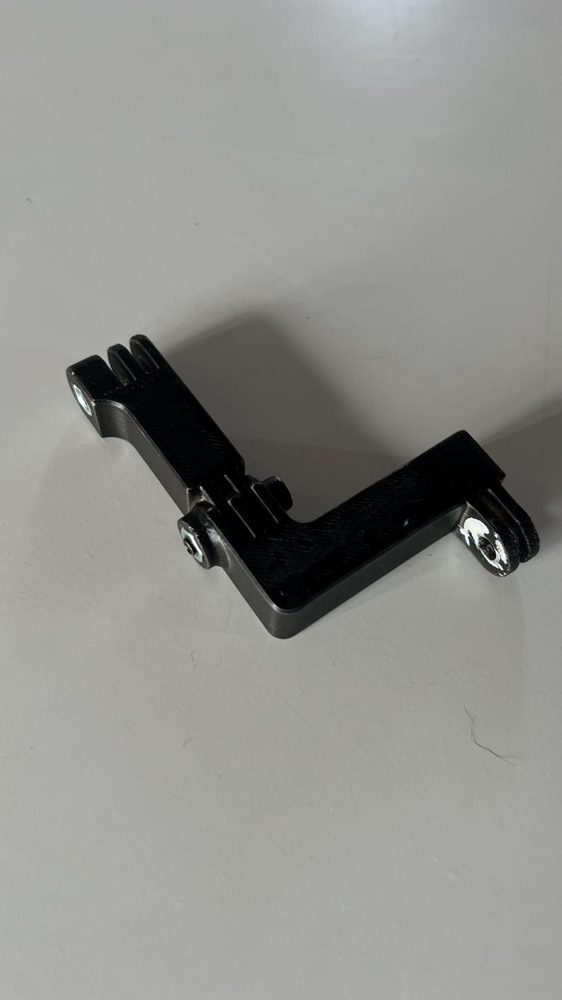 May include: A black, L-shaped metal bracket with two mounting holes and a slotted end. The bracket has a screw in the center and is designed for mounting or securing equipment. The object is on a white surface.
