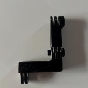 May include: Black plastic L-shaped camera mount with multiple slots and a hinge. The mount is designed to attach to a camera and other accessories. The mount is made of durable plastic.