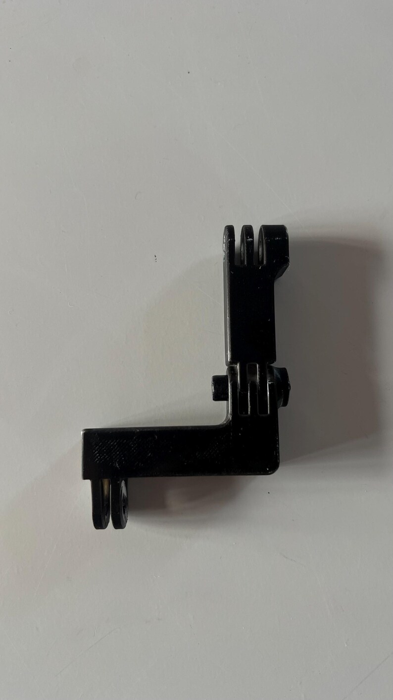 May include: Black plastic L-shaped camera mount with multiple slots and a hinge. The mount is designed to attach to a camera and other accessories. The mount is made of durable plastic.