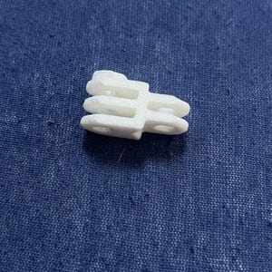 May include: A small, white plastic clip with a unique shape, featuring multiple layers and circular openings. The clip is designed for a specific purpose, likely for securing or connecting items. The object is set against a textured blue background.