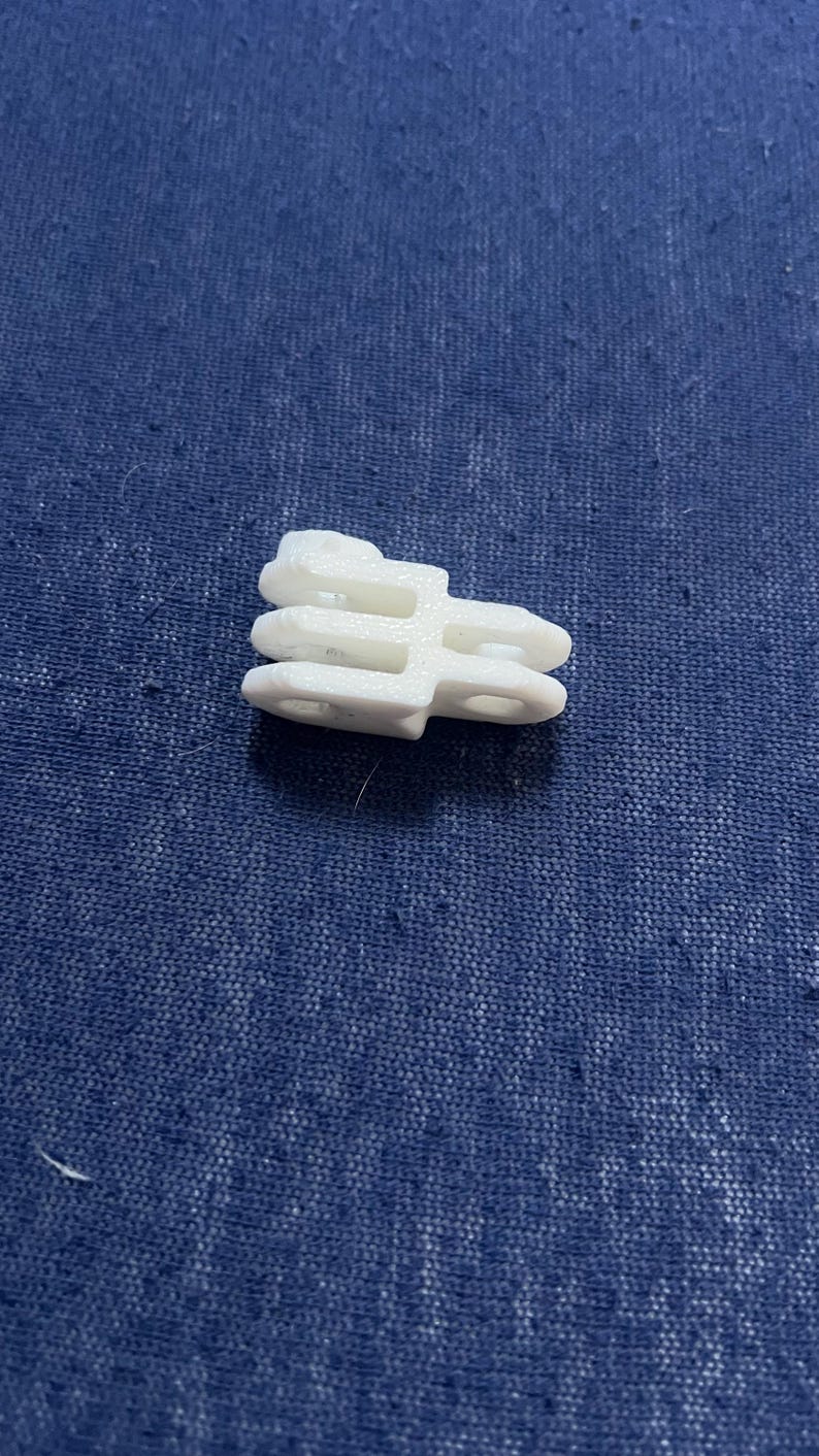 May include: A small, white plastic clip with a unique shape, featuring multiple layers and circular openings. The clip is designed for a specific purpose, likely for securing or connecting items. The object is set against a textured blue background.