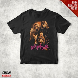 Puede incluir: Camiseta negra con una imagen de estilo vintage de la banda New York Dolls. La imagen utiliza tonos naranja y marrón, con el nombre de la banda en rosa. La camiseta tiene cuello redondo clásico y mangas cortas.