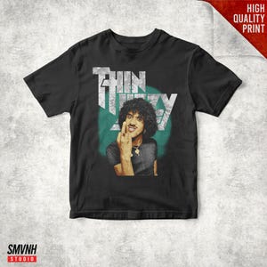 Puede incluir: Camiseta negra con un gráfico de un hombre con pelo rizado, mostrando el dedo medio. El texto "THIN LIZZY" está impreso encima de la imagen en una fuente desgastada, con un círculo verde azulado detrás del hombre. Las palabras "HIGH QUALITY PRINT" están en la esquina superior derecha.