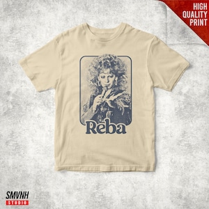 Può includere: T-shirt beige con un'immagine in stile vintage di Reba McEntire. L'immagine è in una cornice rettangolare e include il nome "Reba" in un carattere stilizzato. La maglietta ha maniche corte e scollo tondo.