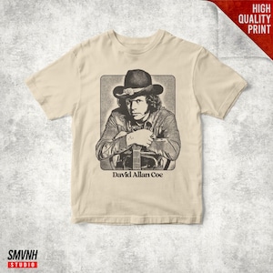 May include: A beige short-sleeve t-shirt featuring a black and white graphic illustration of a person wearing a cowboy hat and holding a guitar. The text David Allan Coe is printed in black lettering directly below the central graphic image.