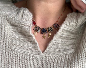 May include: A gold-toned necklace featuring various pendant shapes, including squares with floral and geometric designs, a star, and a lightning bolt. The necklace is worn over a light gray knit sweater.