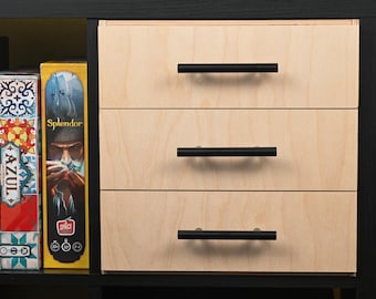 May include: A light-colored wooden storage unit with three horizontal drawers, each featuring a matte black cylindrical handle. To the left, board game boxes labeled Azul and Splendor are visible within a black frame structure.