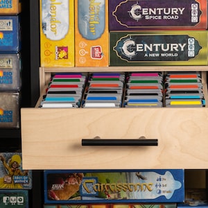 May include: A wooden drawer with a black handle, filled with colorful game cards, sits within a black shelf. The drawer is surrounded by board game boxes, including titles like "Grande Games" and "Century: Spice Road."