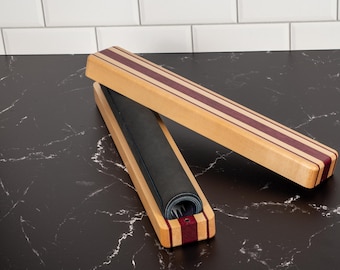 May include: A rectangular wooden storage box with a hinged lid, featuring light tan wood with dark purple striped accents. The box is open, revealing a dark gray, rolled-up flexible mat or sheet stored inside the hollow interior compartment.