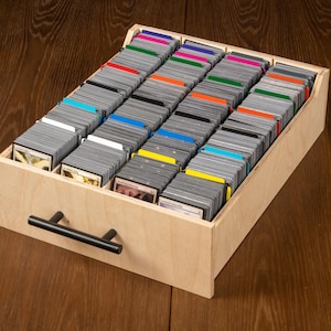 May include: A wooden card storage box filled with rows of trading cards. The cards are organized with colored dividers. The box has a black handle and is sitting on a wooden surface.
