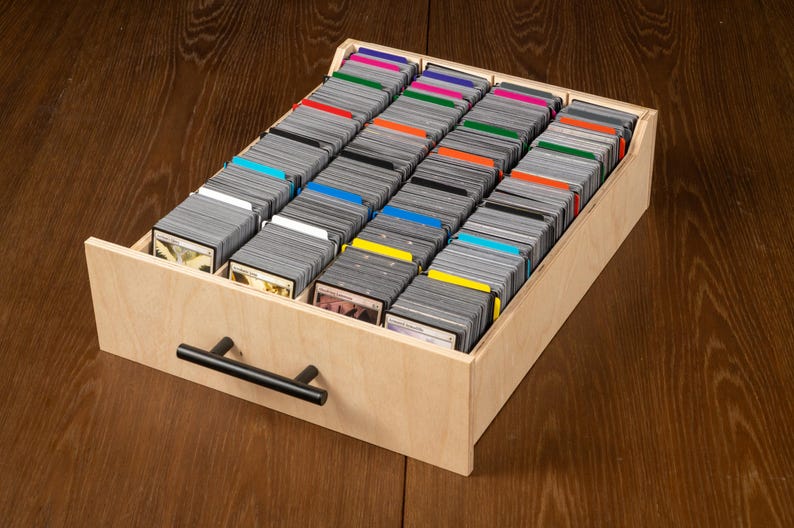 May include: A wooden card storage box filled with rows of trading cards. The cards are organized with colored dividers. The box has a black handle and is sitting on a wooden surface.