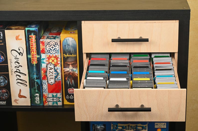 May include: A black and light wood cabinet with two drawers, one open, filled with stacks of colorful cards. Board game boxes line the shelf above. The open drawer reveals a collection of cards, organized by color.