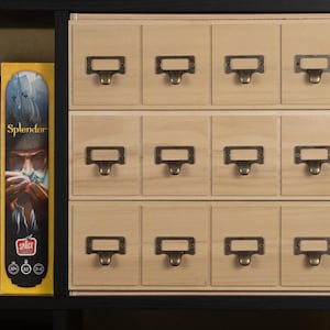 May include: A wooden storage unit with twelve small drawers, each with a metal label holder and pull. The unit is set within a black frame, with board games visible on the left.
