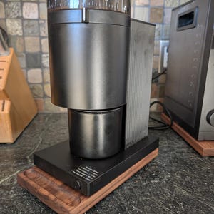 May include: A black electric coffee grinder with a clear top and numbered settings. It sits on a wooden base with a small tray. The grinder is on a dark countertop.
