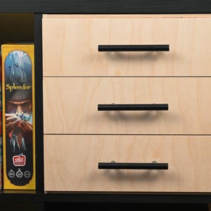 May include: A wooden cabinet with three drawers, each featuring a black handle. The cabinet is constructed with light-colored wood and is set within a black frame. Board game boxes are visible on the left side of the cabinet.