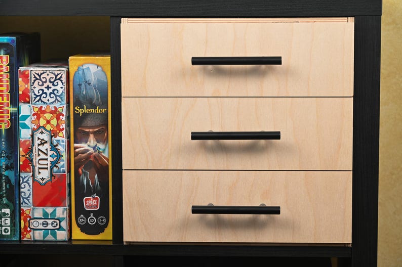 May include: A wooden cabinet with three drawers, each featuring a black handle. The cabinet is constructed with light-colored wood and is set within a black frame. Board game boxes are visible on the left side of the cabinet.