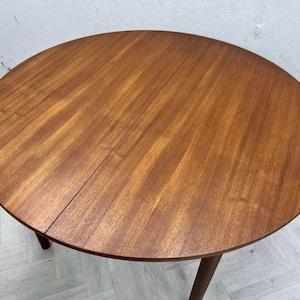 May include: A round wooden dining table with a warm brown finish and a visible grain pattern. The tabletop features a central seam indicating an extendable design, supported by tapered wooden legs that extend downward from the circular surface.