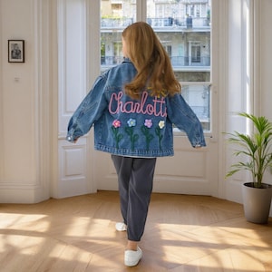 May include: A blue denim jacket with the name "Charlott" embroidered in pink script. Below the name are embroidered flowers in various colours. The jacket is worn with grey trousers and white shoes.