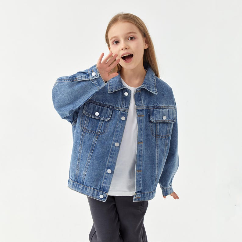 May include: A light blue denim jacket with white buttons and two chest pockets. The jacket is worn over a white t-shirt. The model is wearing dark gray pants.