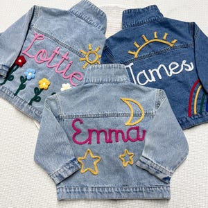 May include: Three denim jackets with embroidered details. One jacket has the name "Lottie" in pink, with flower and sun designs. Another has "James" with a sun and rainbow. The third jacket has "Emma" with a moon and stars.