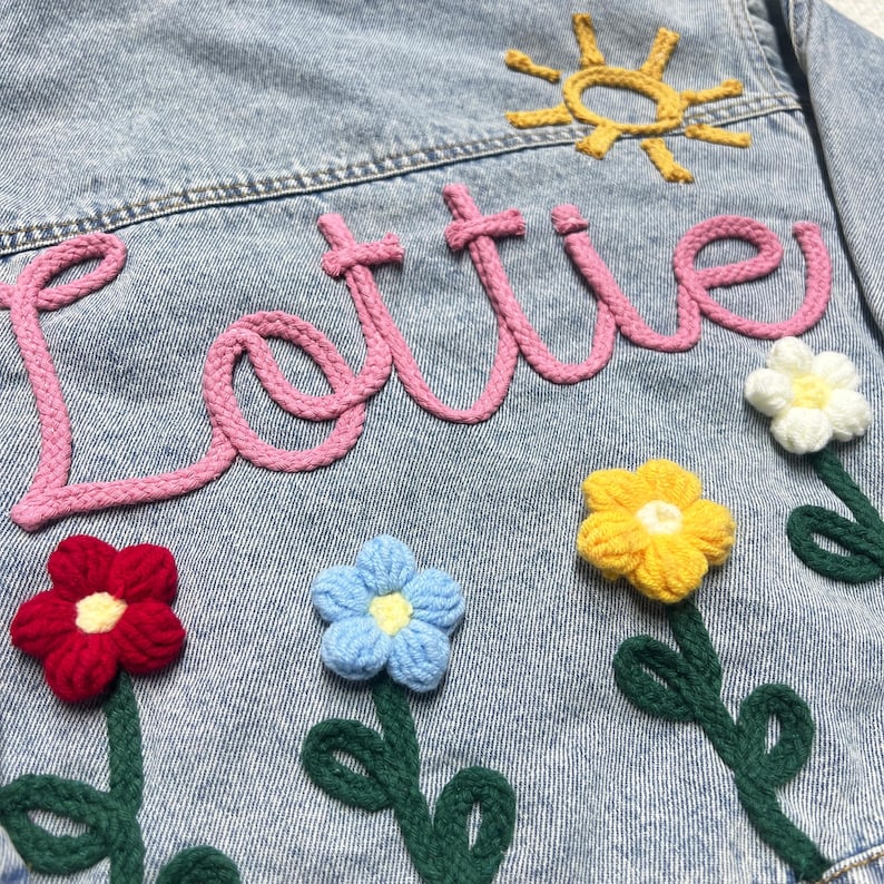May include: A light blue denim jacket with the name "Lottie" embroidered in pink yarn. The jacket is decorated with crocheted flowers in red, yellow, blue, and white.