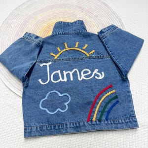 May include: A blue denim jacket with embroidered details. The back of the jacket features a yellow sun, a blue cloud, a rainbow, and the name "James" in white thread.