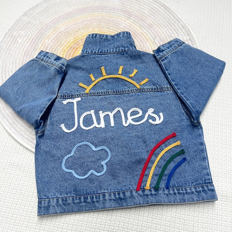 May include: A blue denim jacket with embroidered details. The back of the jacket features a yellow sun, a blue cloud, a rainbow, and the name "James" in white thread.
