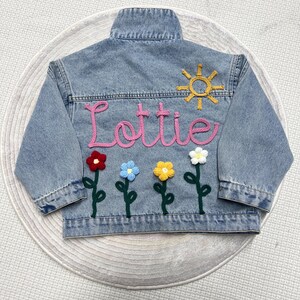 May include: A light blue denim jacket with the name "Lottie" embroidered in pink yarn. The jacket is decorated with a yellow sun and five crocheted flowers in red, blue, yellow, and white.