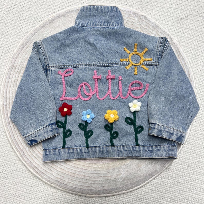 May include: A light blue denim jacket with the name "Lottie" embroidered in pink yarn. The jacket is decorated with a yellow sun and five crocheted flowers in red, blue, yellow, and white.