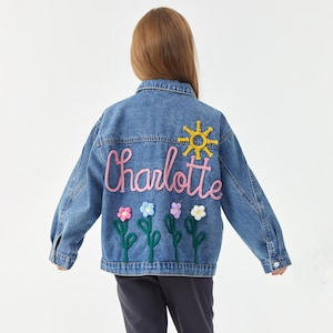 May include: A denim jacket with the name "Charlotte" embroidered in pink script, a yellow sun, and colorful flower appliqu&eacute;s. The jacket is a light blue wash and has a classic button-up design.