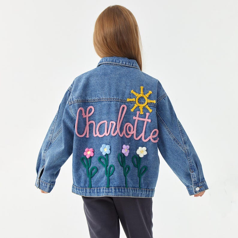 May include: A denim jacket with the name "Charlotte" embroidered in pink script, a yellow sun, and colorful flower appliqu&eacute;s. The jacket is a light blue wash and has a classic button-up design.
