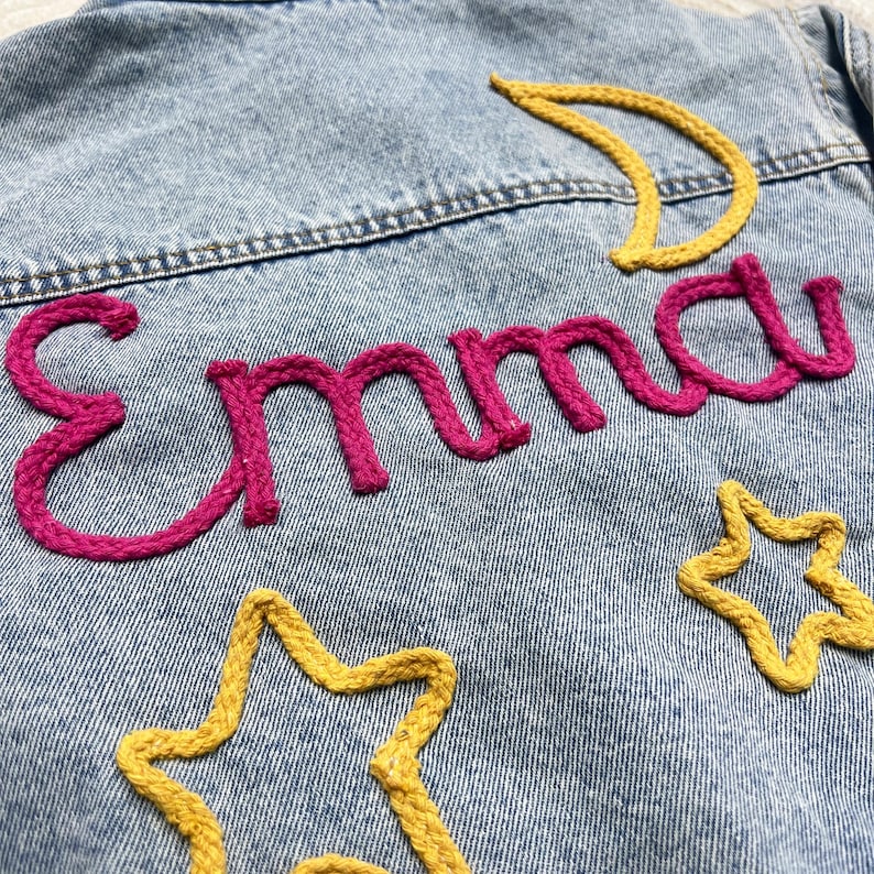 May include: A denim jacket with the name "Emma" embroidered in pink yarn. The jacket also features yellow yarn stars and a crescent moon.