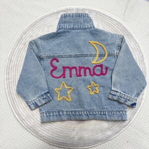 May include: A light blue denim jacket with the name "Emma" embroidered in pink yarn. The jacket also features a yellow crescent moon and two yellow stars.