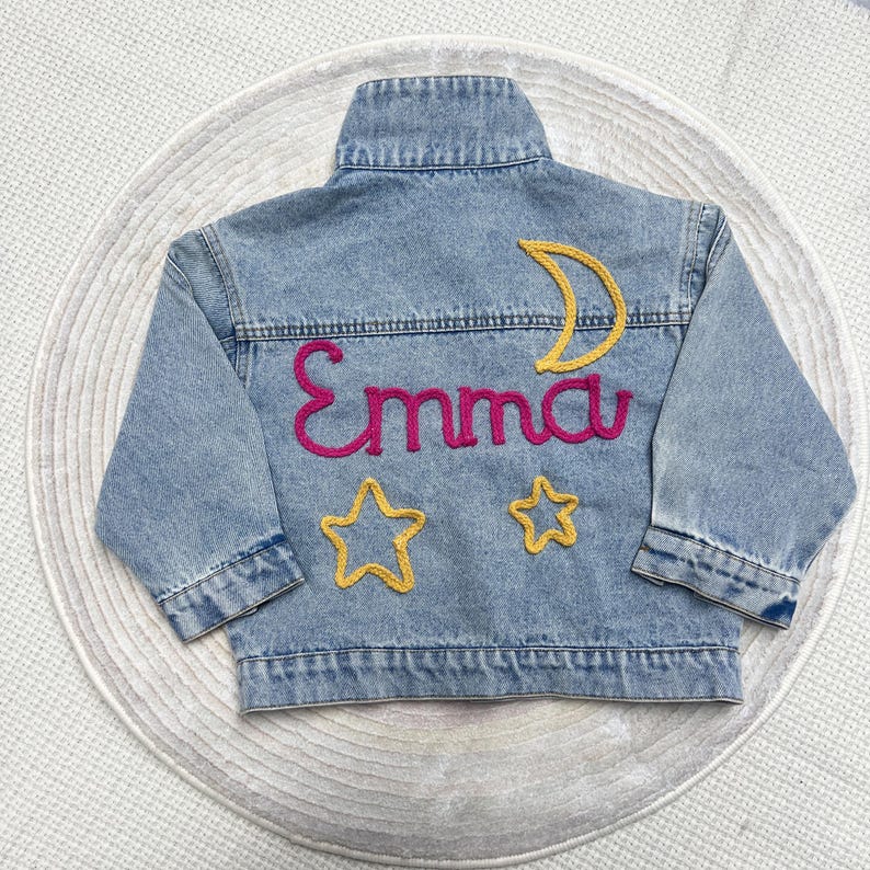 May include: A light blue denim jacket with the name "Emma" embroidered in pink yarn. The jacket also features a yellow crescent moon and two yellow stars.