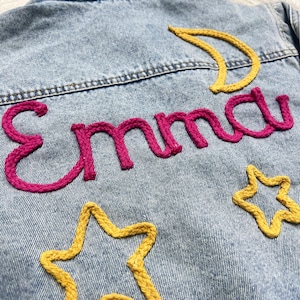 May include: Light blue denim jacket with embroidered details. The name "Emma" is stitched in bright pink, with yellow stars and a crescent moon. The jacket has a classic design.