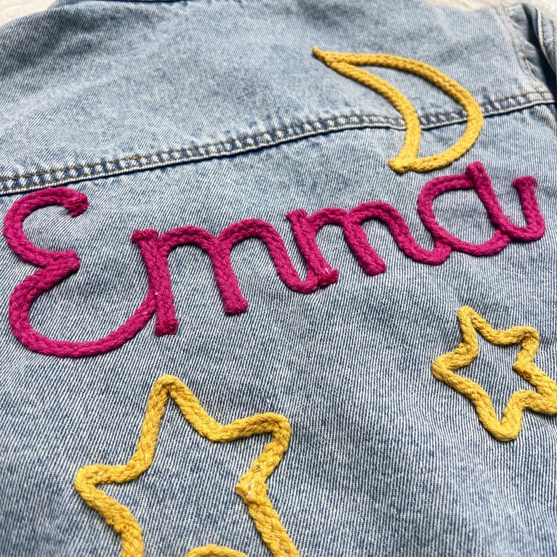 May include: Light blue denim jacket with embroidered details. The name "Emma" is stitched in bright pink, with yellow stars and a crescent moon. The jacket has a classic design.