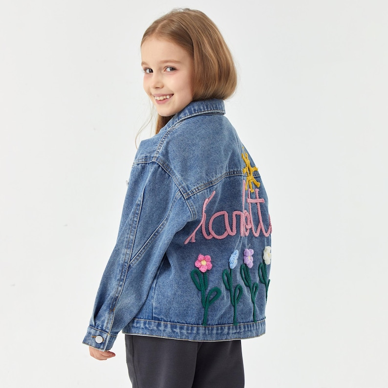 May include: A blue denim jacket with colorful embroidered flowers and the word "Lanett" on the back. The jacket has a classic collar and button closures. The flowers are in shades of pink, blue, and white.