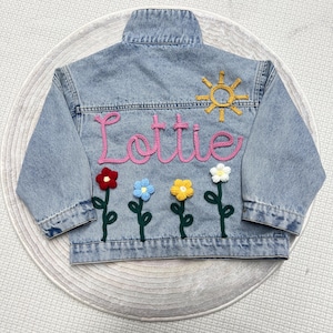 May include: Light blue denim jacket with the name "Lottie" in pink yarn. The back of the jacket features a yellow sun and embroidered flowers in red, blue, yellow, and white. A stylish and personalized children's jacket.