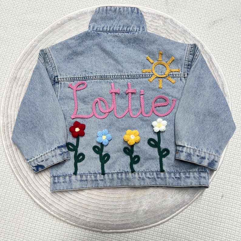 May include: Light blue denim jacket with the name "Lottie" in pink yarn. The back of the jacket features a yellow sun and embroidered flowers in red, blue, yellow, and white. A stylish and personalized children's jacket.