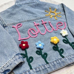 May include: Pale blue denim jacket featuring the name "Lottie" in pink yarn. The back is adorned with crocheted flowers in red, blue, yellow, and white, and a yellow sun. A distinctive, personalised children's jacket.