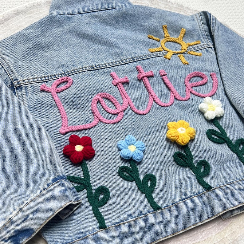 May include: Light blue denim jacket with the name "Lottie" in pink yarn. The back is decorated with crocheted flowers in red, blue, yellow, and white, and a yellow sun. A unique and personalized children's jacket.