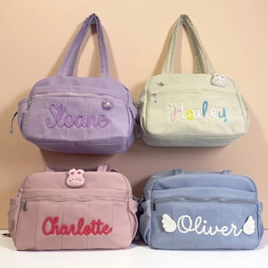 May include: Four pastel-coloured baby bags, each personalised with a name. The bags are lavender, light green, pink, and blue, with embroidered names and decorative details. They feature a quilted design and zipped pockets. The bags have handles for carrying.